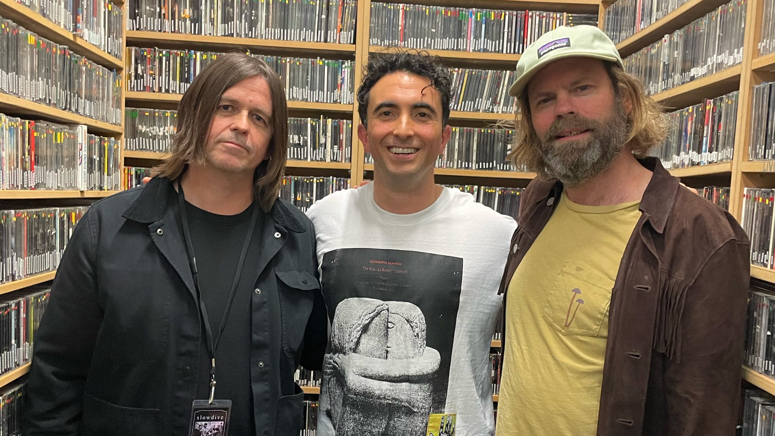 Slowdive members Neil and Simon stand next in the FBi CD library room, with presetner Al Grigg in between them. All three are facing the camera for a photo.