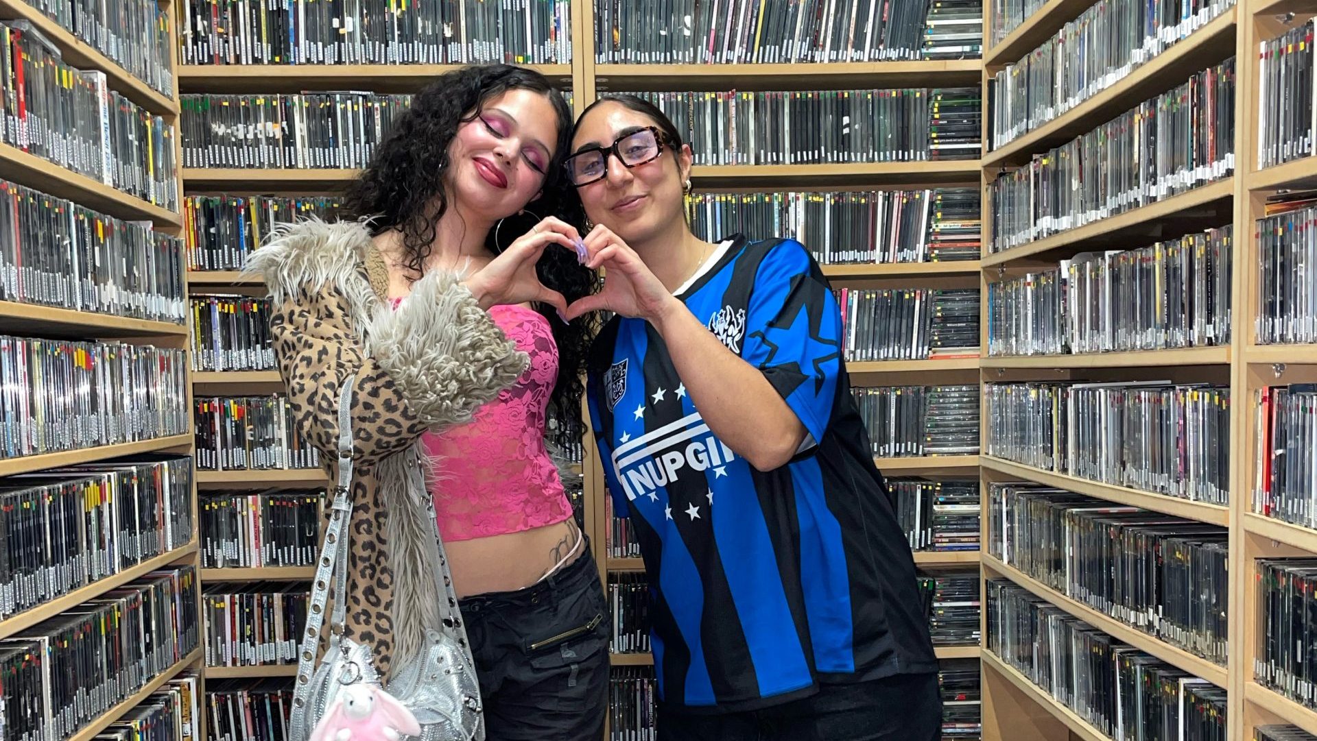 Naz and Cherry Chola stand in the music library. They are making a love heart shape with their hands.